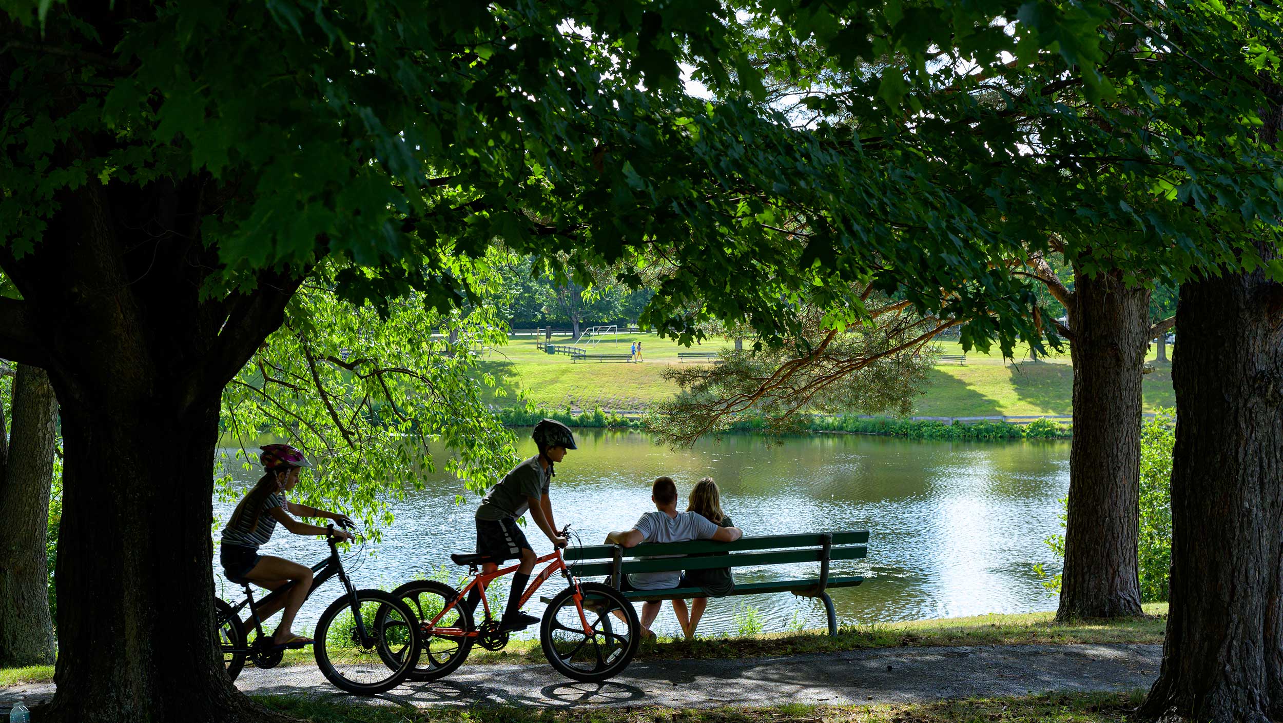 Two kids riding bicycles, near two people sitting on a park bench